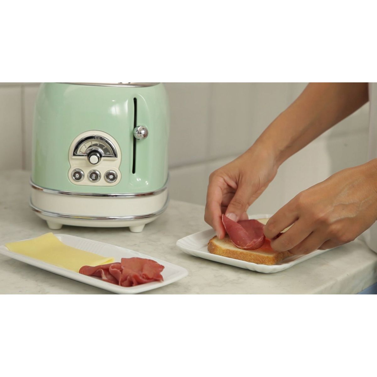 Eine Person belegt Toast mit Schinken neben einem mintgrünen Toaster.