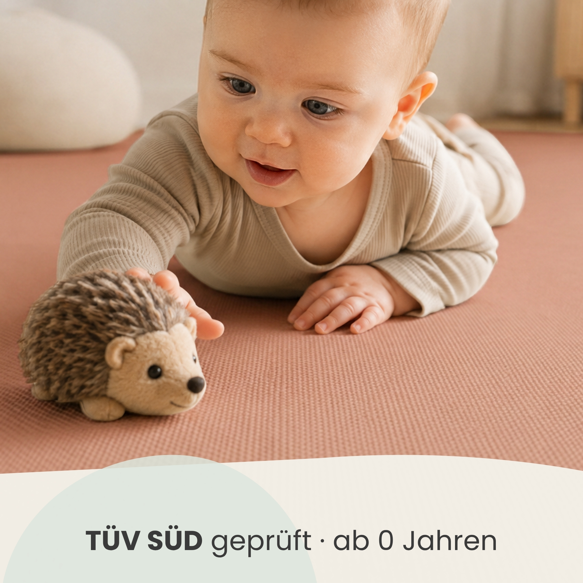 Ein Baby liegt auf einer rosa Matte und greift nach einem Plüschigel. Unten steht 'TÜV SÜD geprüft'.