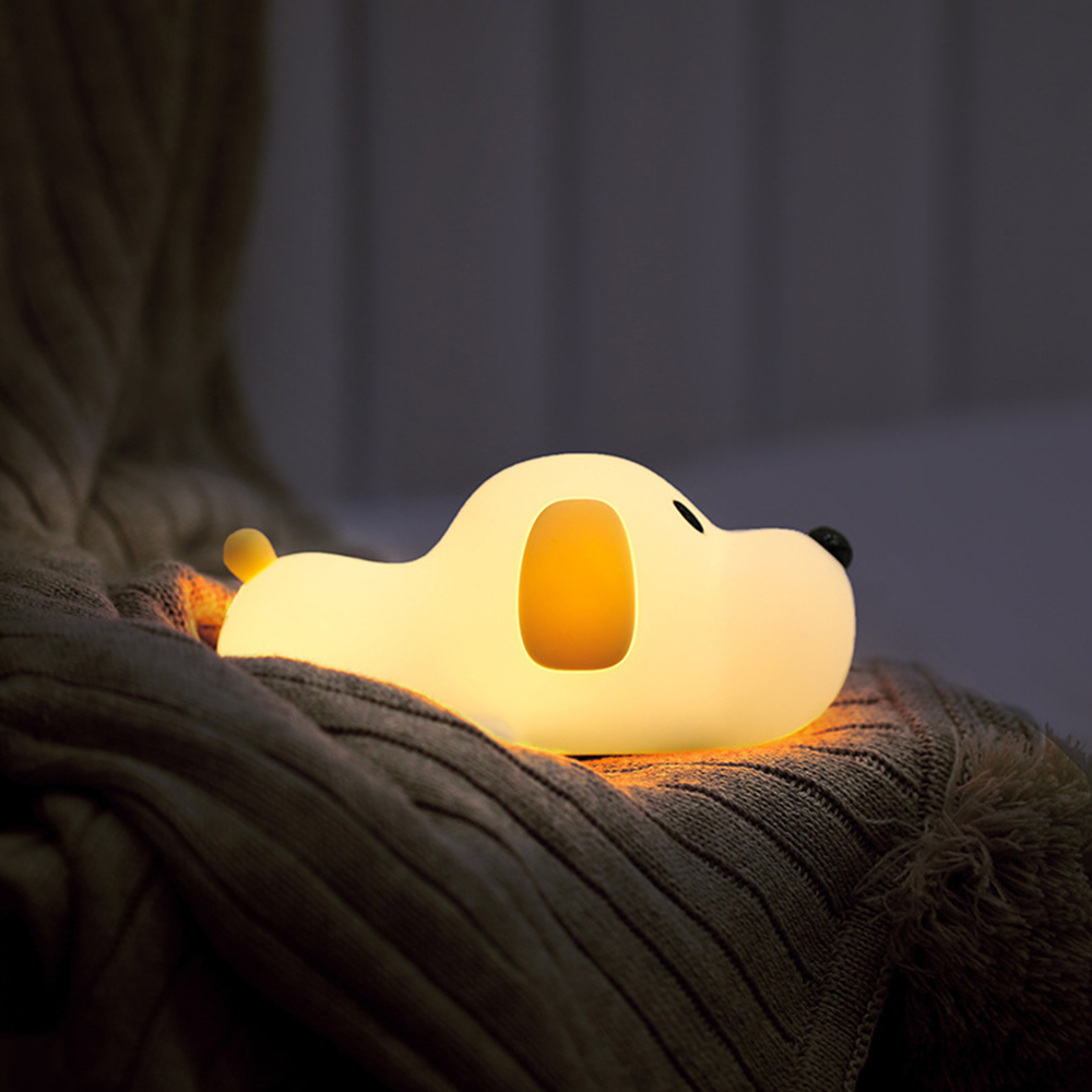 Eine kleine, warm leuchtende, hundeförmige Nachtlampe ruht auf einer weichen, strukturierten Decke in einem dunklen Raum.