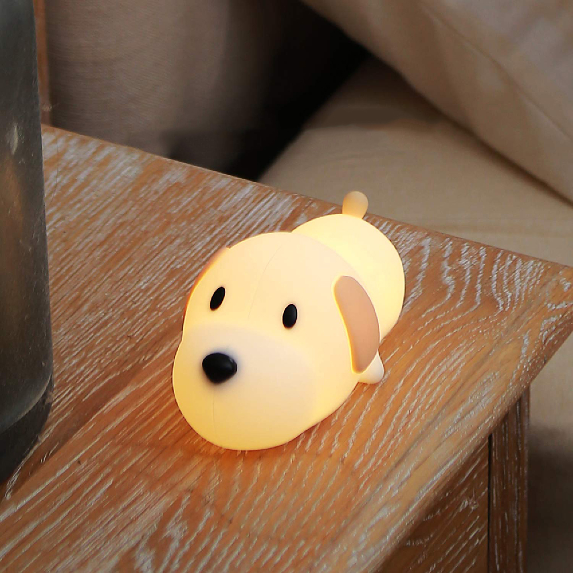 Eine kleine, leuchtende Hundelampe aus Silikon steht auf einem Holztisch in einem ruhigen Raum.