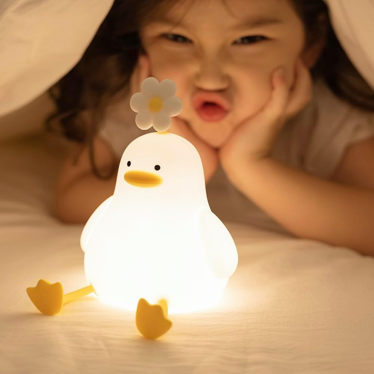 Ein kleines Mädchen schaut lächelnd unter einer Decke hervor, während eine leuchtende Entenlampe im Vordergrund steht.