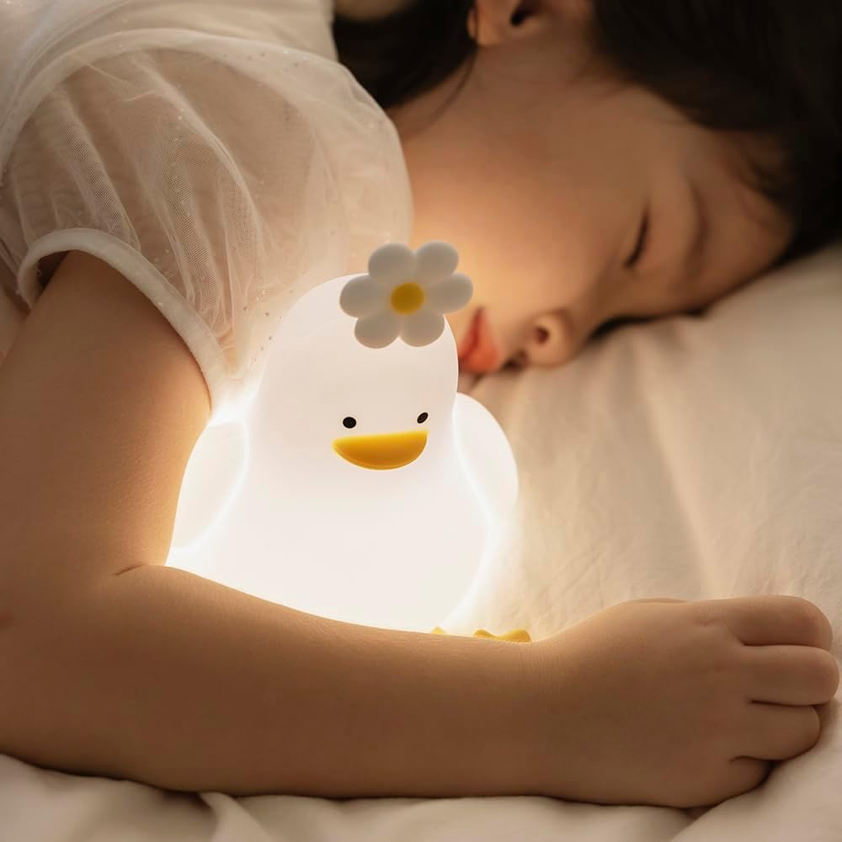 Ein kleines Kind schläft friedlich in einem weißen Bett neben einem leuchtenden, weichen Enten-Nachtlicht mit Blume.