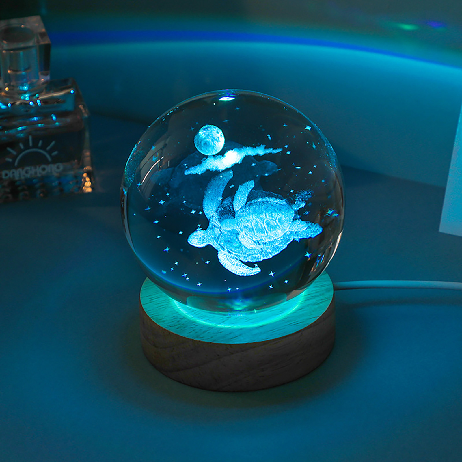Eine leuchtende Kristallkugel auf einem Holzsockel zeigt eine Schildkröte zwischen Sternen und Mond im blauen Licht.