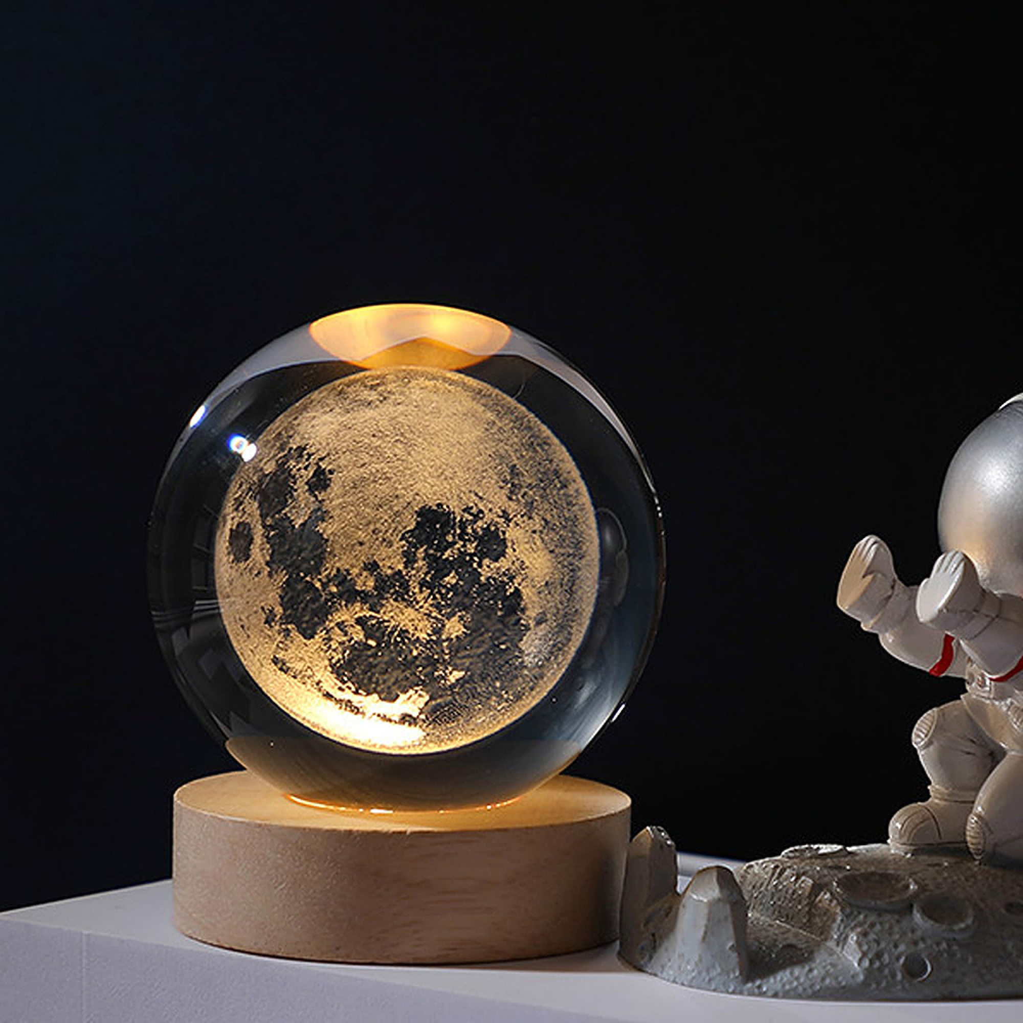Eine leuchtende Kristallkugel mit dem Mond auf einem Holzsockel neben einer kleinen Astronautenfigur vor schwarzem Hintergrund.