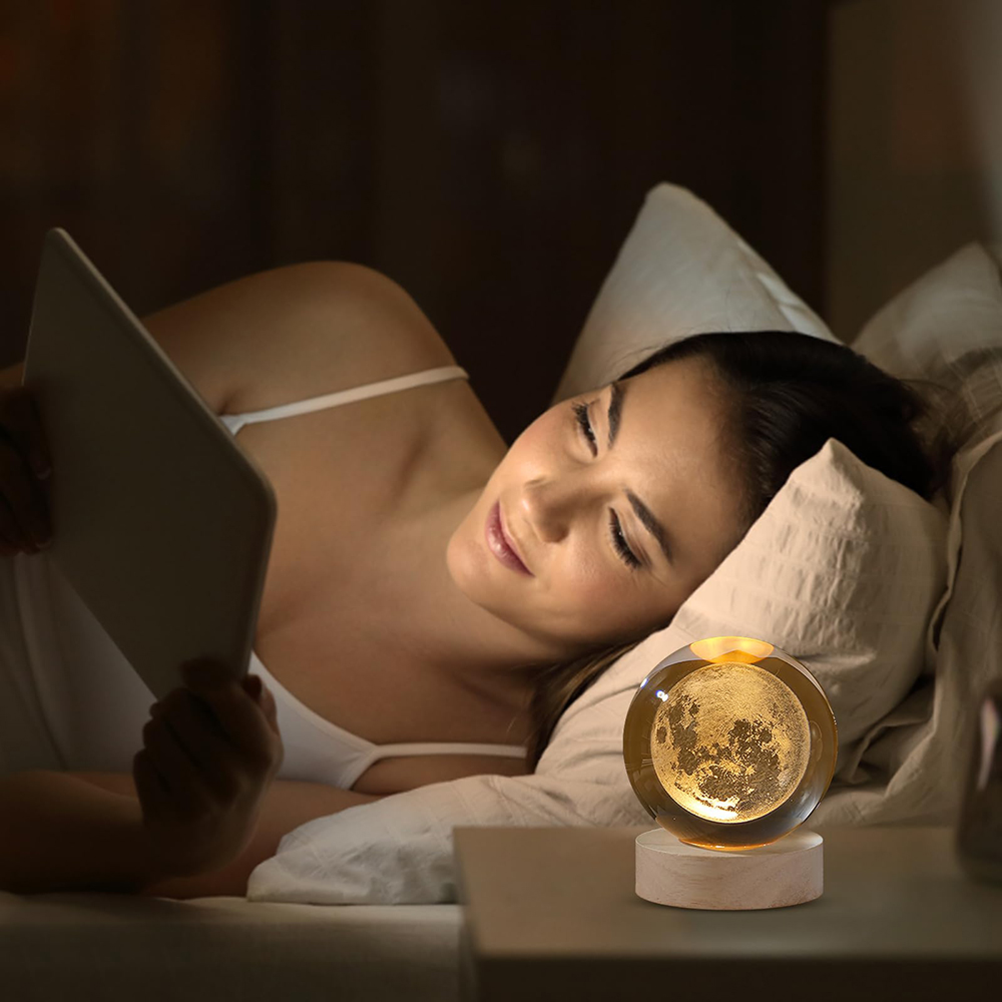 Eine Frau liegt im Bett und schaut auf ein Tablet, während eine glühende Mondlampe auf dem Nachttisch steht.