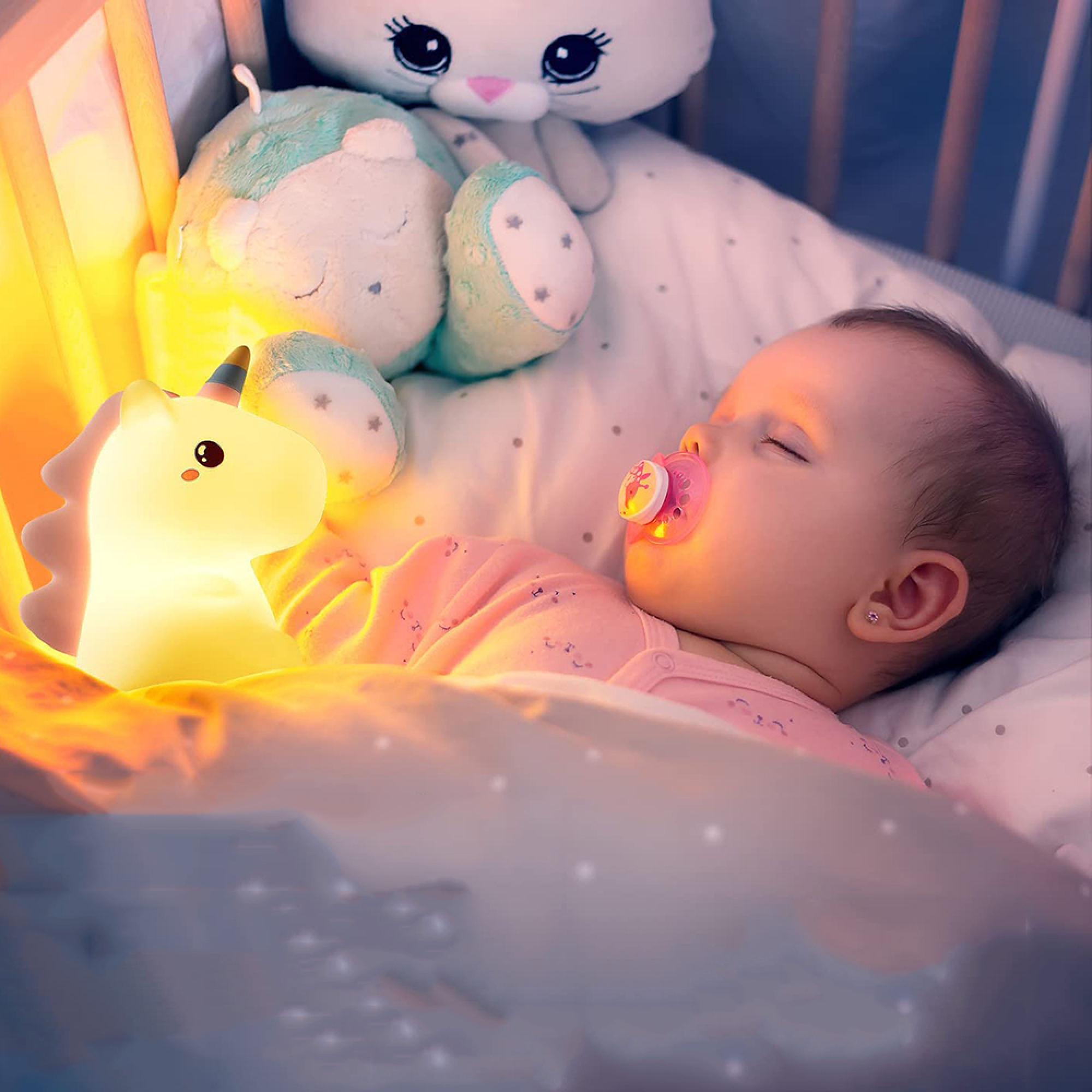 Ein Baby schläft mit einem Schnuller im Bett, umgeben von Plüschtieren und einem Einhorn-Nachtlicht.