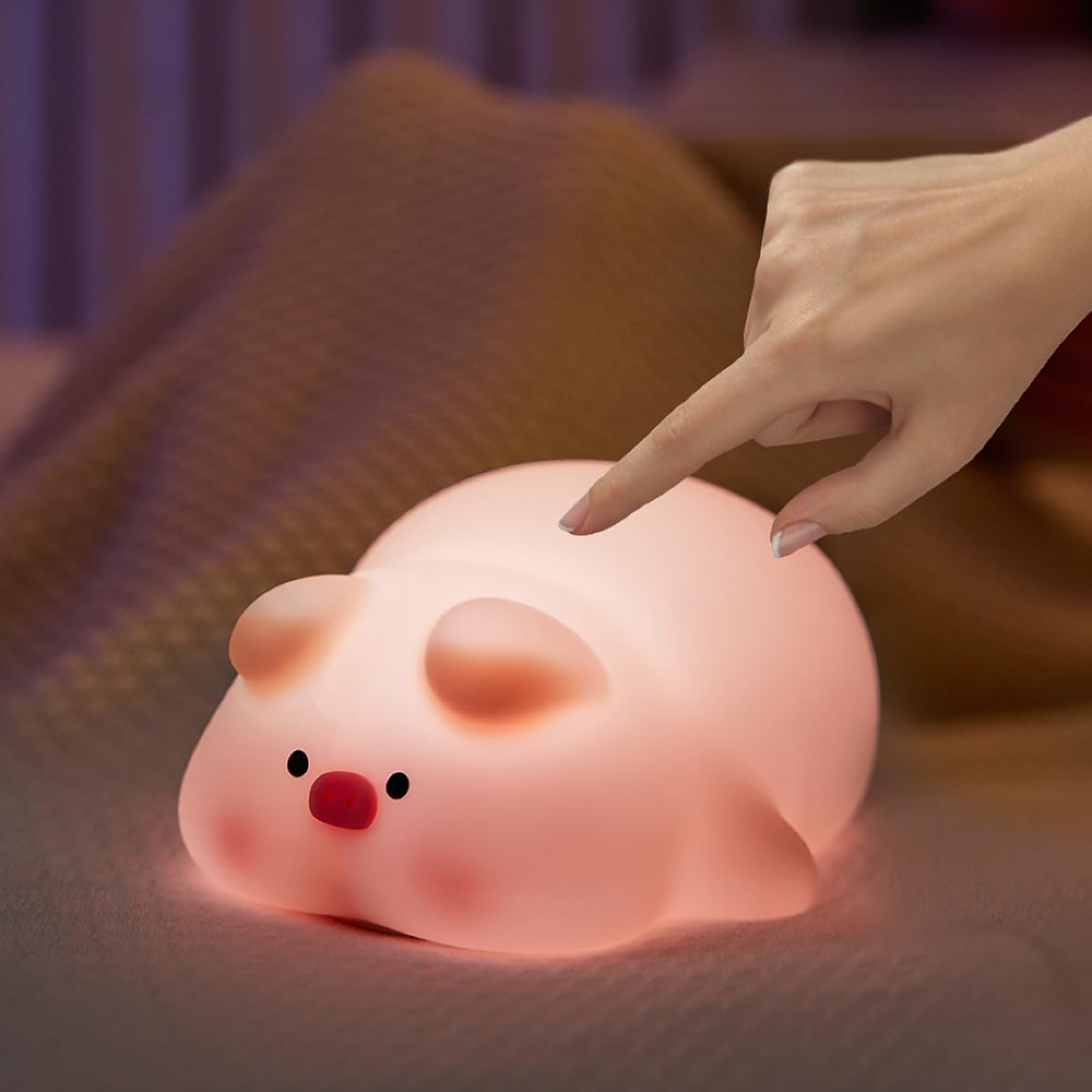 Eine Hand tippt auf ein kleines, leuchtendes Schweinchen-Nachtlicht auf einer Bettdecke.