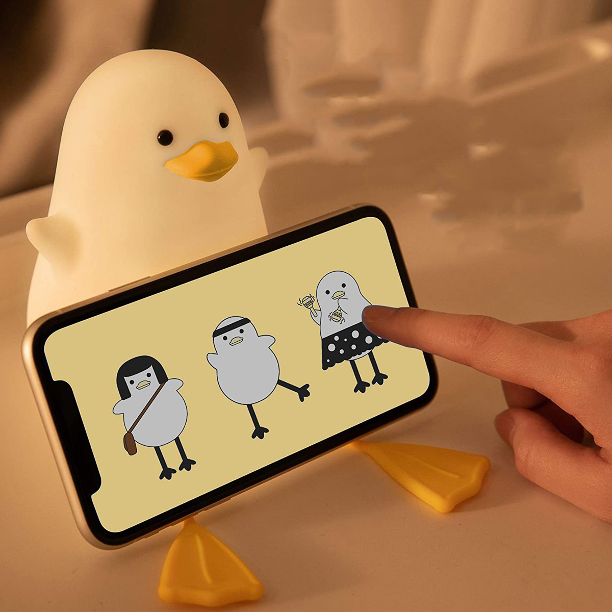 Eine Enten-Nachtleuchte hält ein Smartphone, auf dessen Bildschirm eine Hand tippt und drei Cartoon-Enten zu sehen sind.