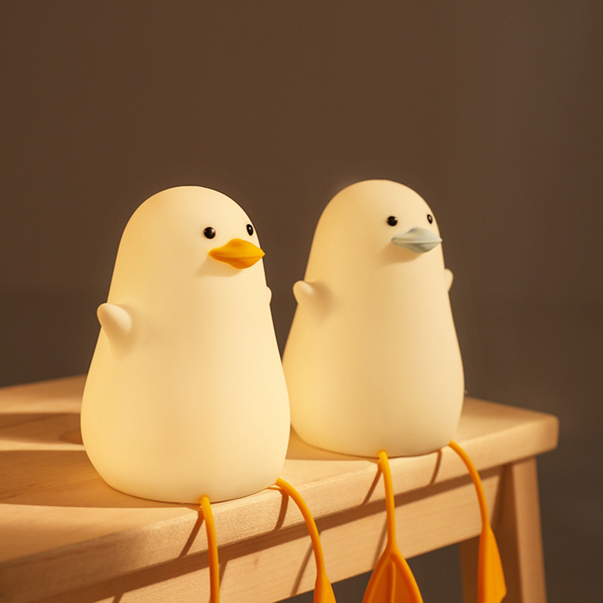 Zwei weiße Enten-Nachtlichter mit langen orangen Beinen sitzen auf einem Holzstuhl vor dunklem Hintergrund.