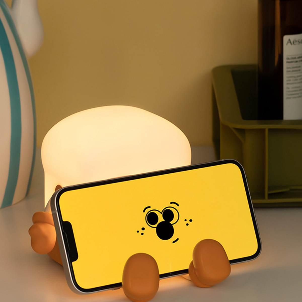 Eine niedliche Handyhalterung mit Lampe zeigt ein Smartphone mit einem lustigen Gesicht auf dem gelben Display.