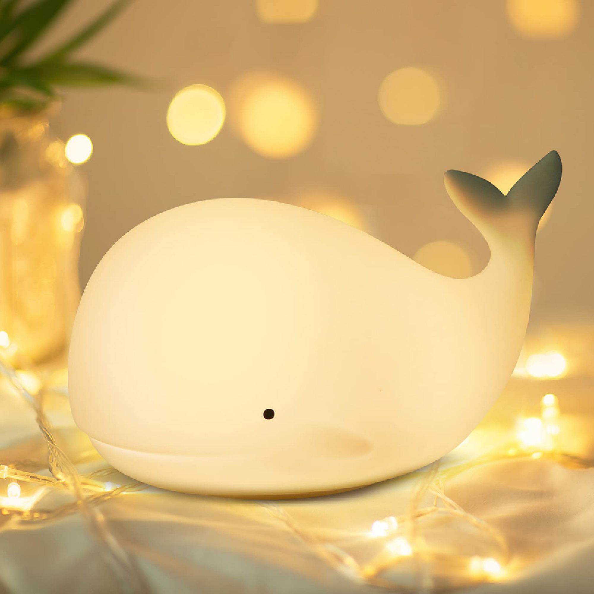Eine weiße Wal-Nachtlampe leuchtet auf einem Tisch mit funkelnden Lichterketten im Hintergrund.
