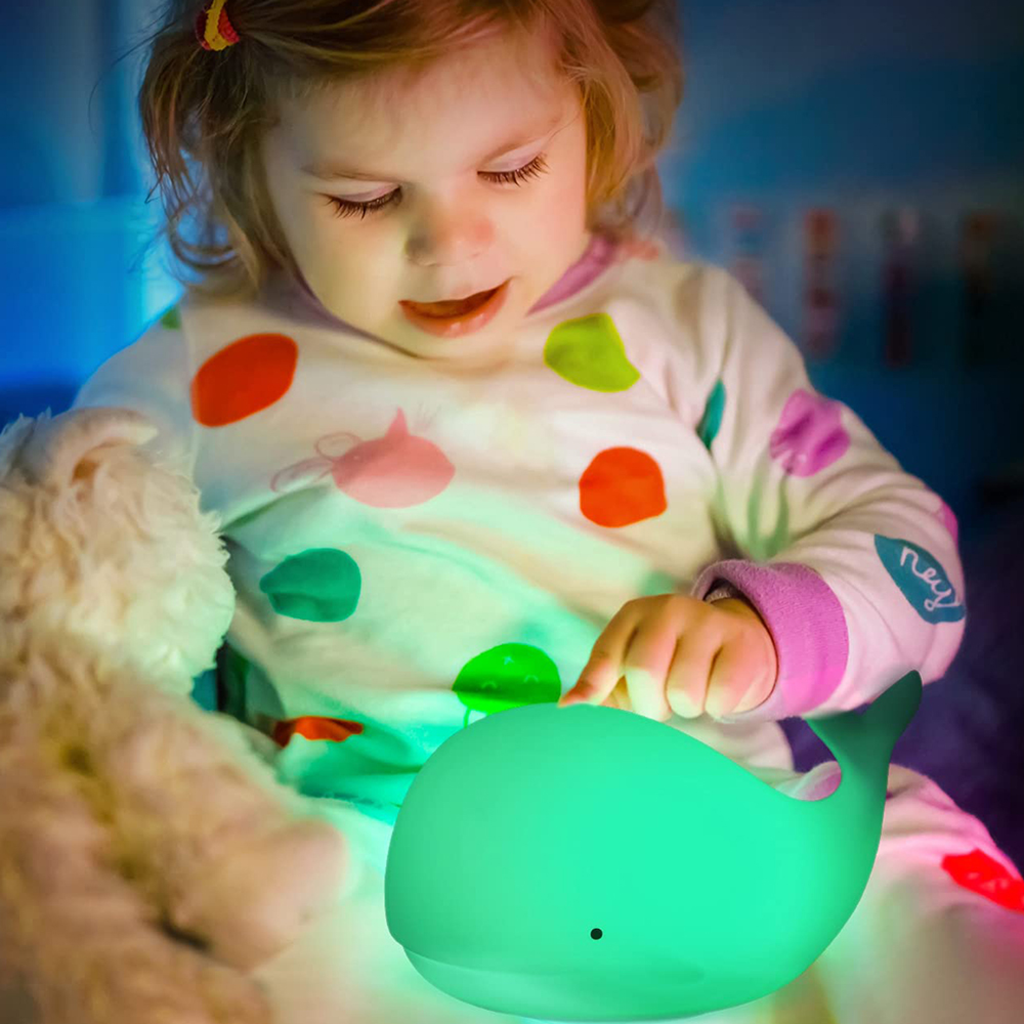 Kleines Mädchen in gepunktetem Schlafanzug tippt auf eine grüne, leuchtende Wal-Lampe neben einem Stofftier.