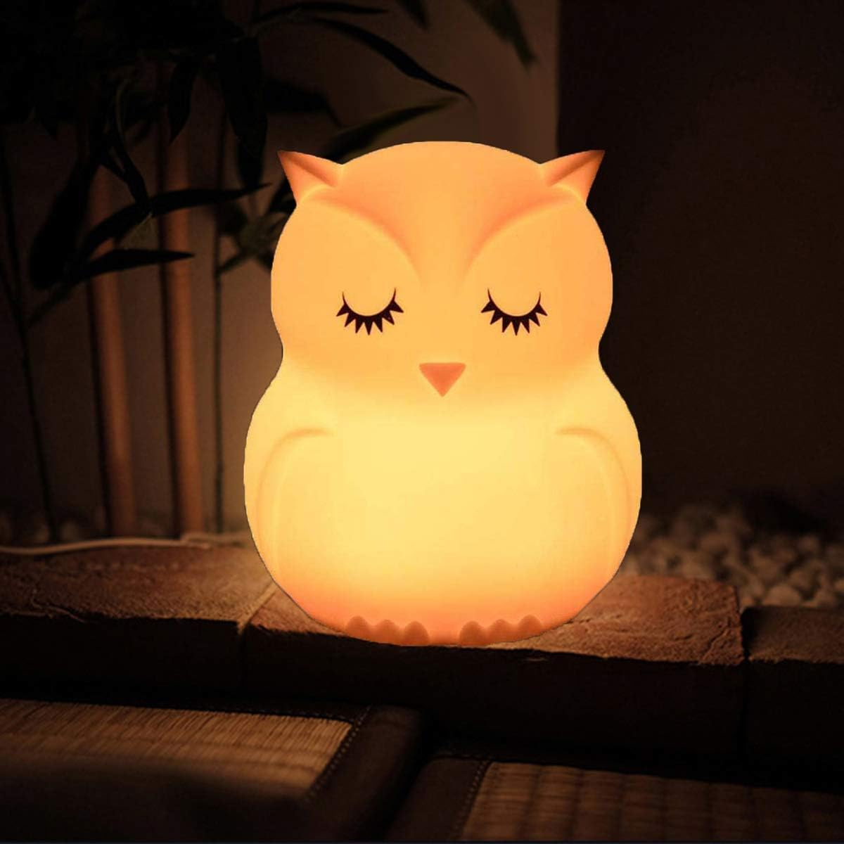 Eine niedliche Eulen-Nachtlampe leuchtet warm orange auf einem dunklen Regal vor grünen Pflanzen.