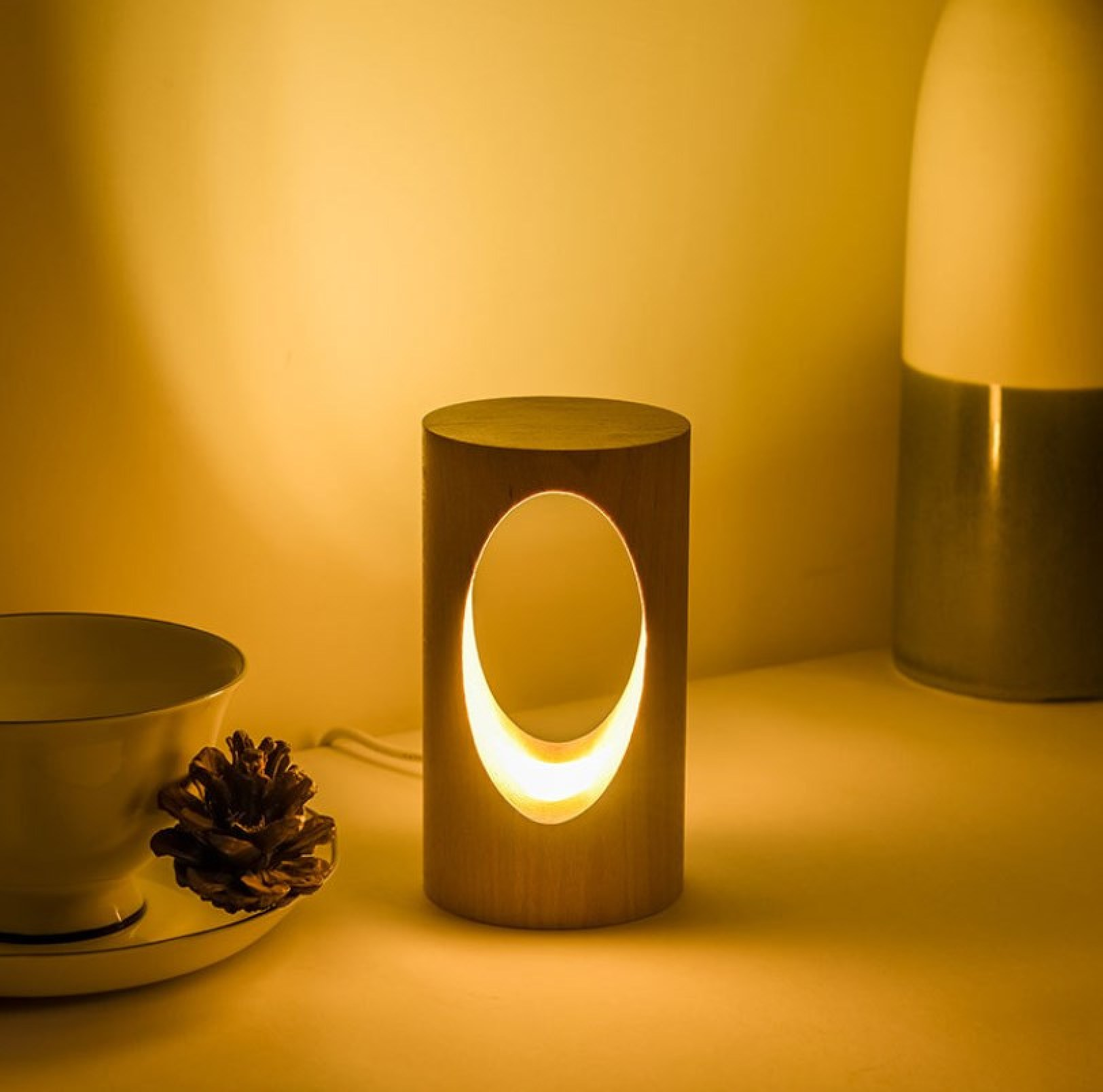 Eine beleuchtete zylindrische Holzlampe mit ovalem Ausschnitt steht neben Tasse, Tannenzapfen und Vase.
