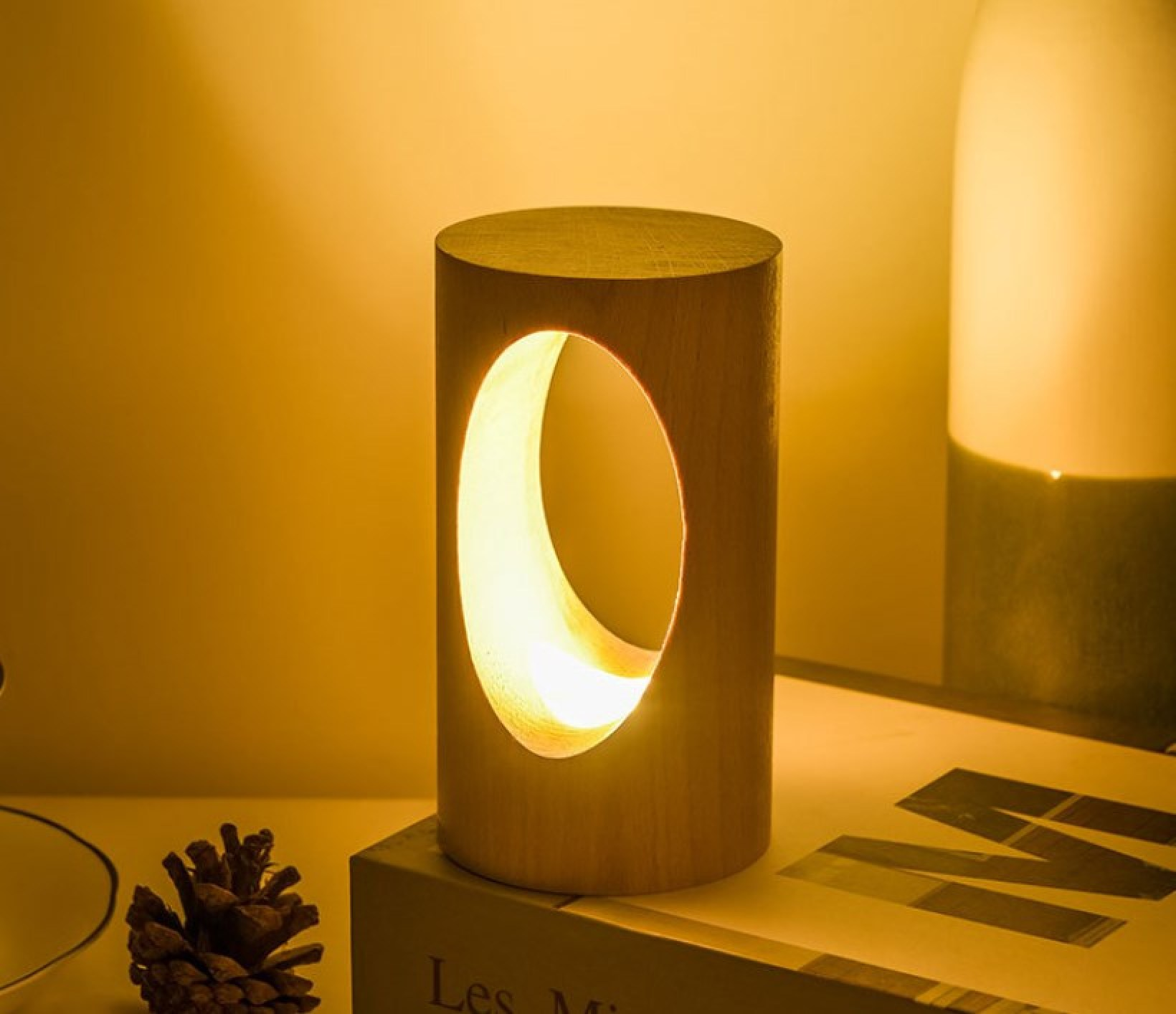 Eine zylindrische Holzlampe mit ovalem Ausschnitt steht auf einem Buch neben einem Tannenzapfen in warmem Licht.