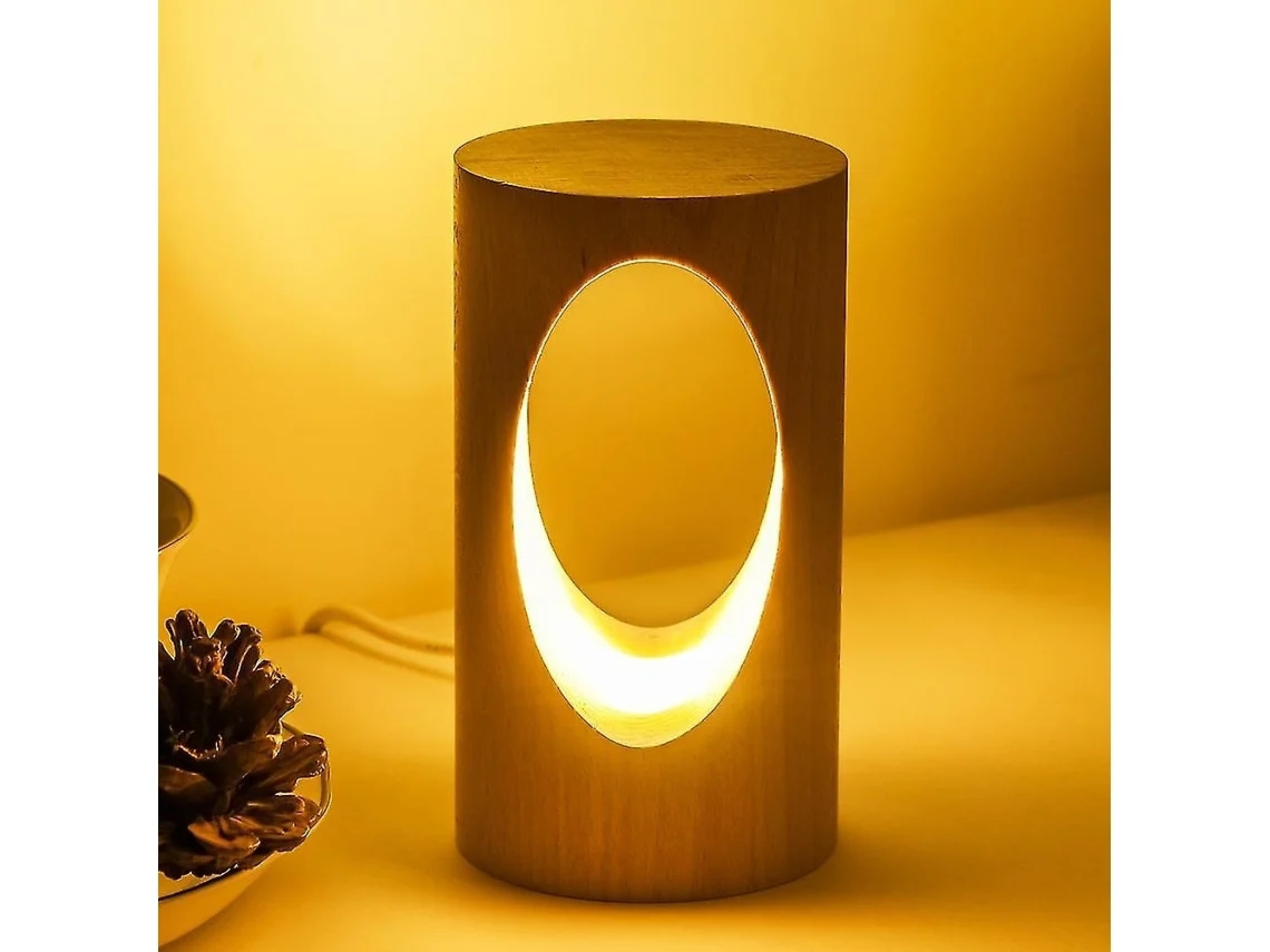 Eine hölzerne Zylinderlampe mit ovalem Ausschnitt leuchtet warm vor einer beigefarbenen Wand, daneben ein Zapfen.