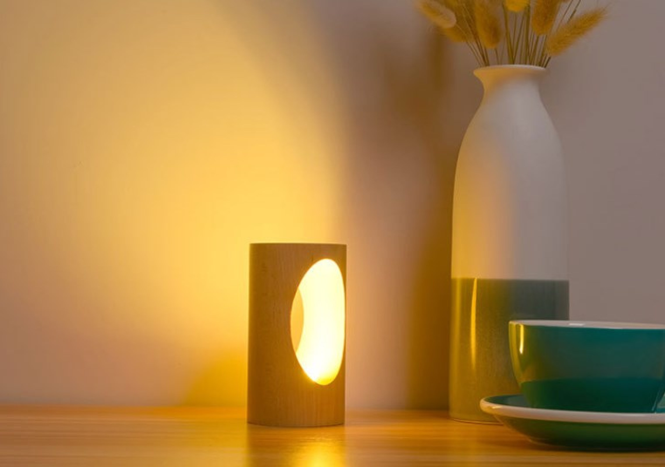Eine beleuchtete Holz-Zylinderlampe wirft ein warmes Licht an eine Wand neben einer weißen Vase mit getrocknetem Gras.