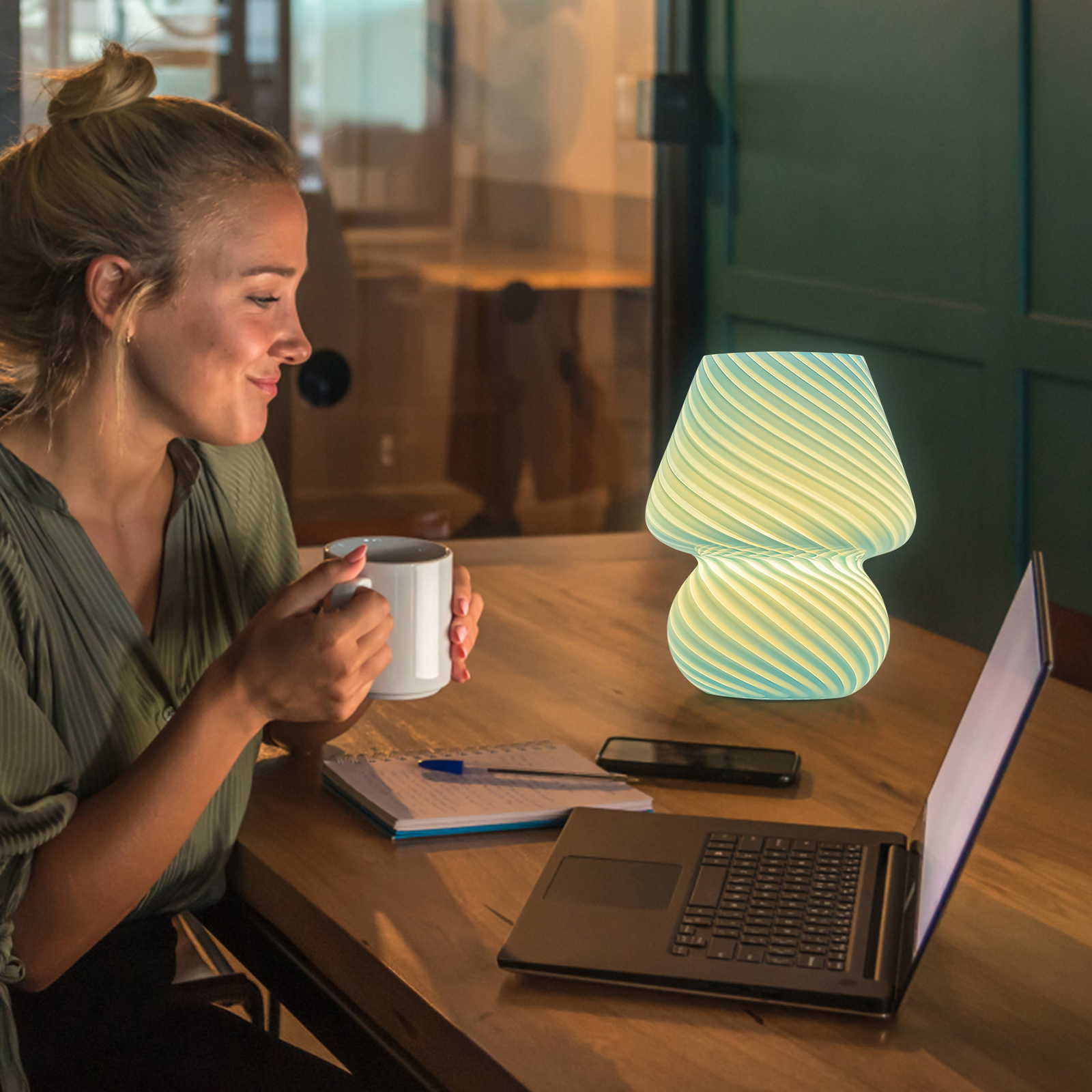 Eine blonde Frau hält eine weiße Tasse an einem Holztisch mit Laptop, leuchtender Lampe, Notizbuch und Telefon.