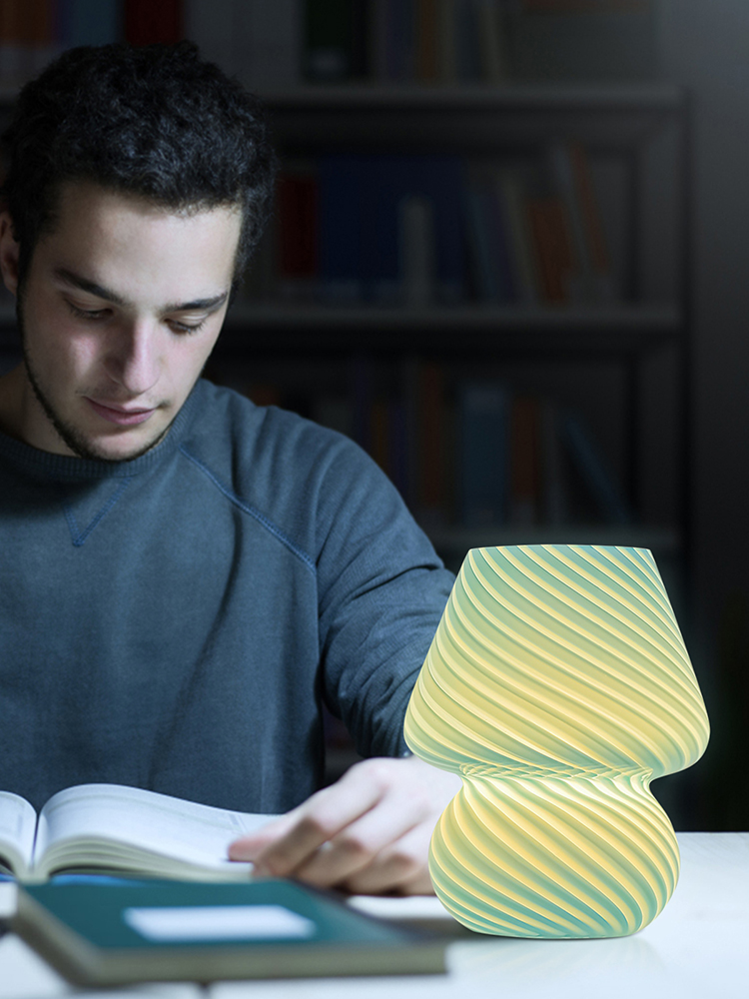 Ein junger Mann liest nachts an einem Schreibtisch neben einer leuchtenden Lampe ein Buch.