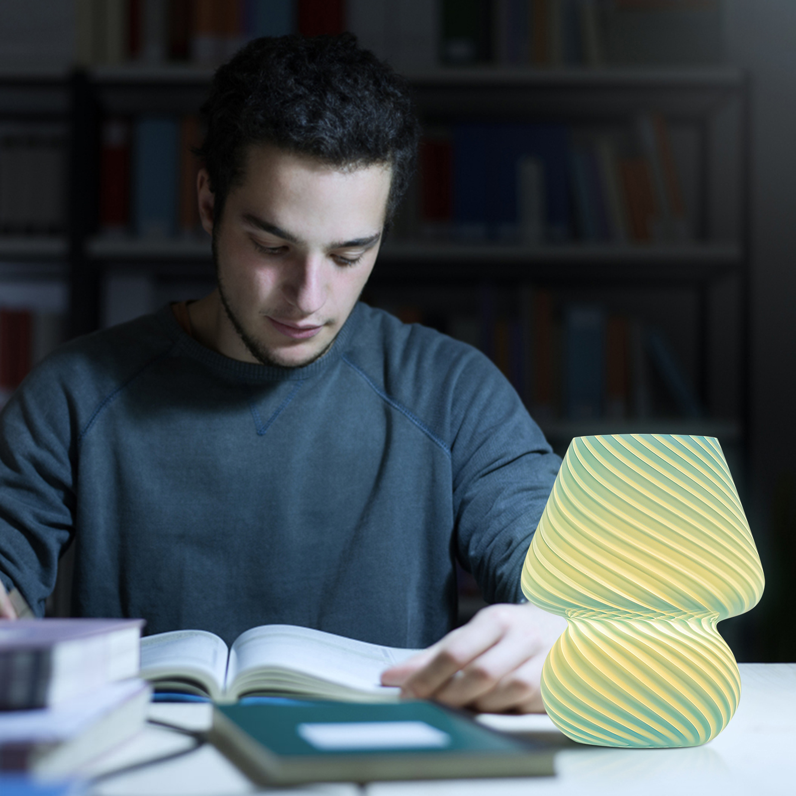 Ein junger Mann liest nachts ein Buch an einem Schreibtisch neben einer glühenden grünen Lampe.