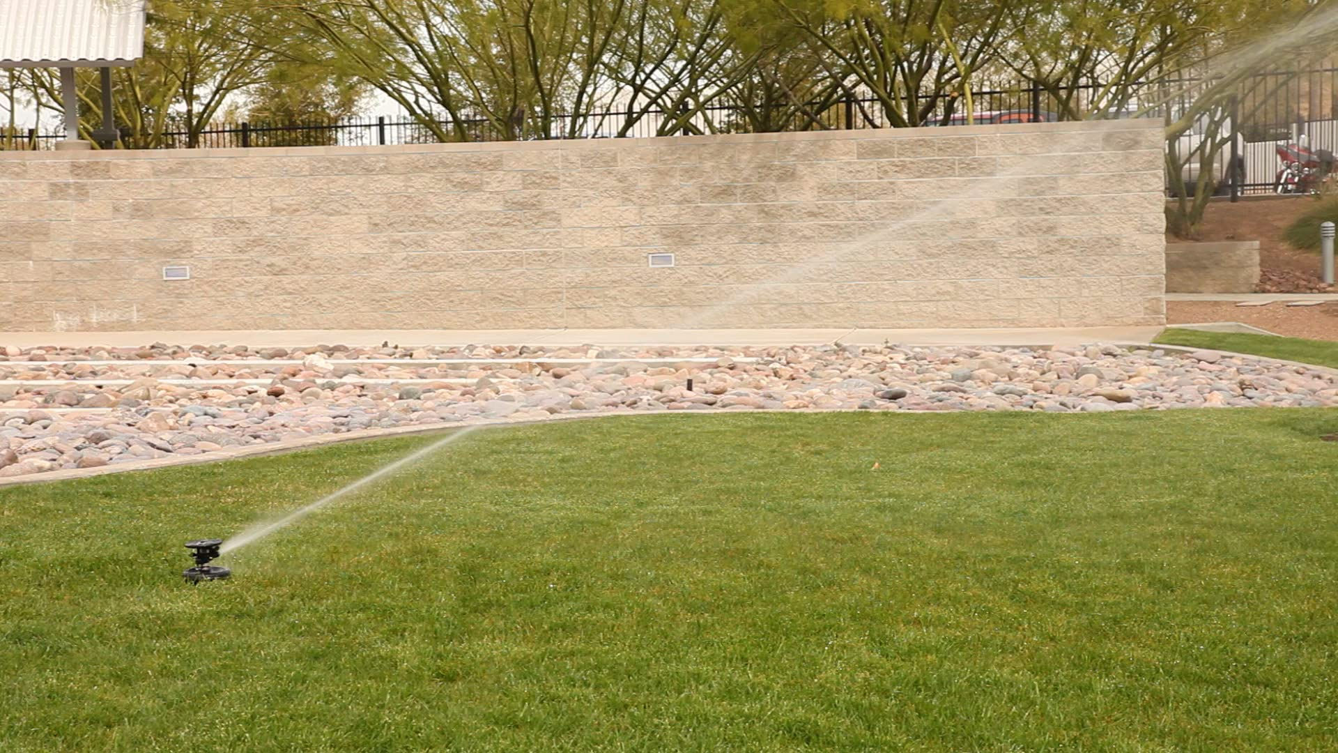 Ein Rasensprenger bewässert eine grüne Wiese vor einer grauen Steinmauer mit Büschen und Bäumen im Hintergrund.