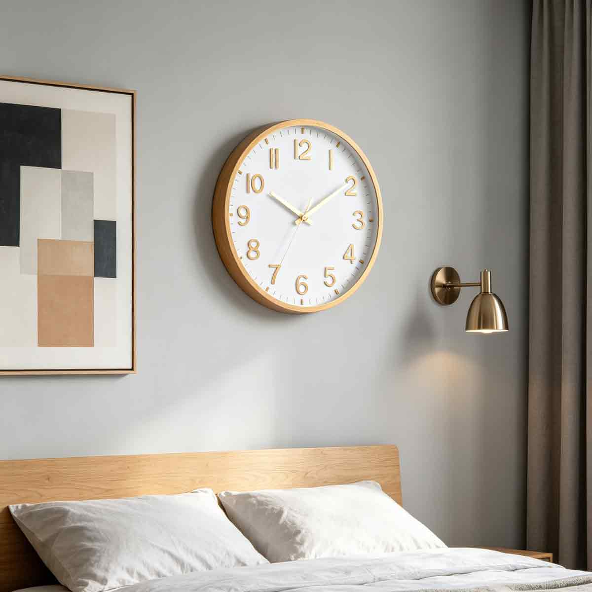 Eine Uhr und ein gerahmtes Bild hängen an einer grauen Wand über einem Holzbett mit weißen Kissen und einer goldenen Lampe.