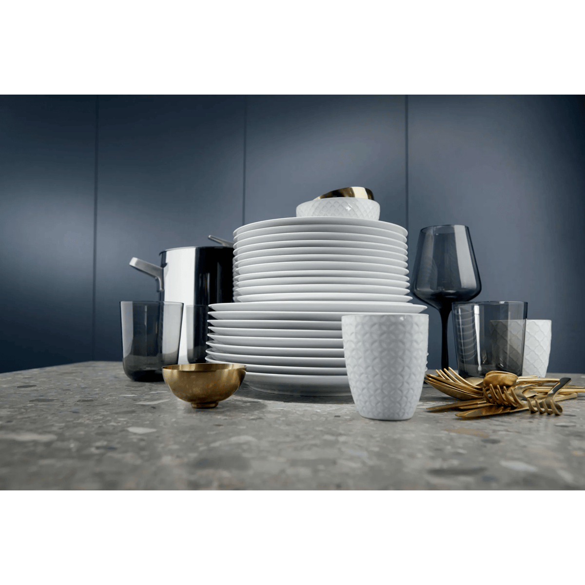 Stack of white plates and cups, gold cutlery, and glasses on a gray granite countertop.