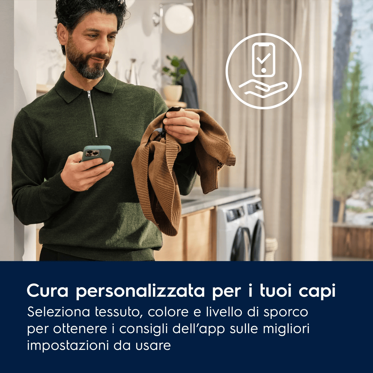 Un uomo con camicia verde controlla l'etichetta di un maglione marrone tenendo il telefono, con lavatrici sullo sfondo.