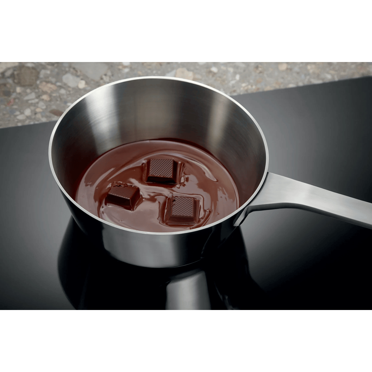 A metal saucepan on a black stovetop contains melted chocolate and three solid chocolate squares.