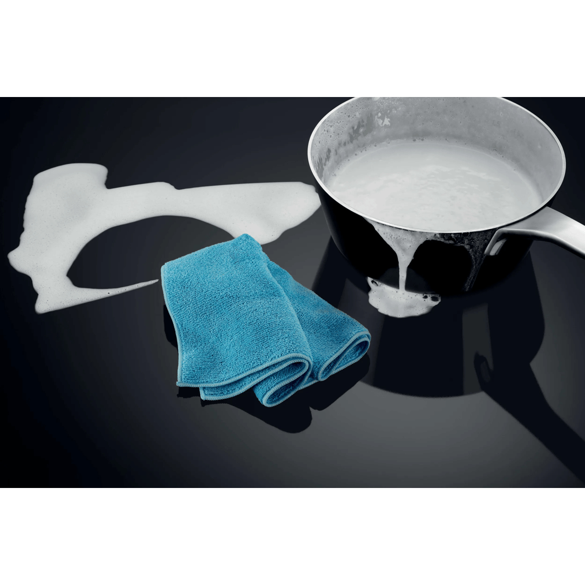 A pot with foam, a blue cloth, and foam traces on a black stovetop.