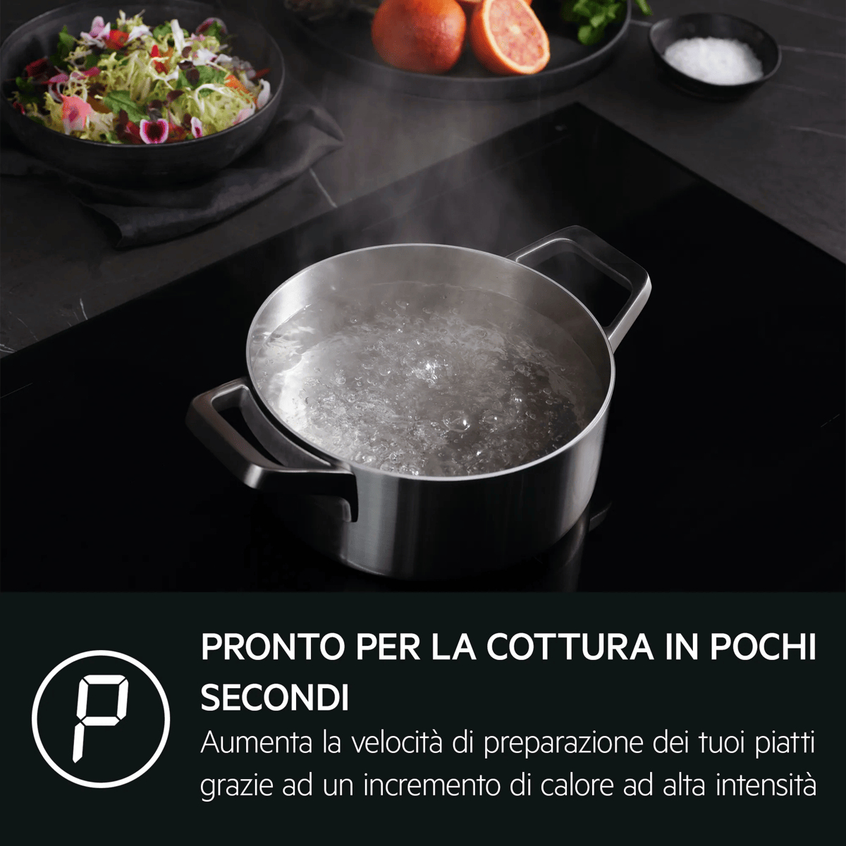 Pentola in acciaio inox con acqua bollente su piano cottura a induzione nero, con insalata e arance sullo sfondo.