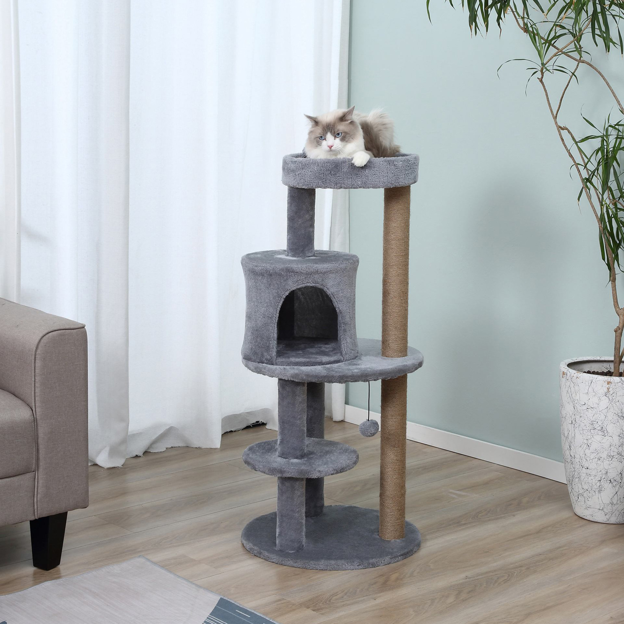Un gatto soffice riposa su un tiragraffi grigio in un soggiorno con pianta e divano.