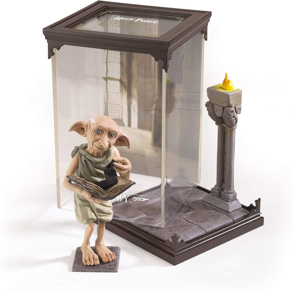 Una figura de Dobby de Harry Potter sostiene un libro frente a una vitrina de cristal con base de piedra y antorcha.