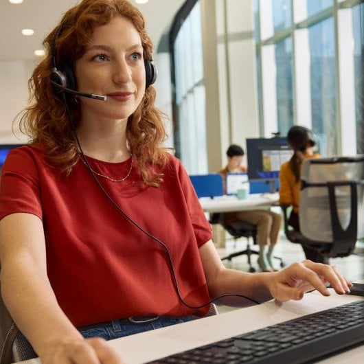 Donna dai capelli rossi con cuffie e top rosso digita al computer in un ufficio luminoso.