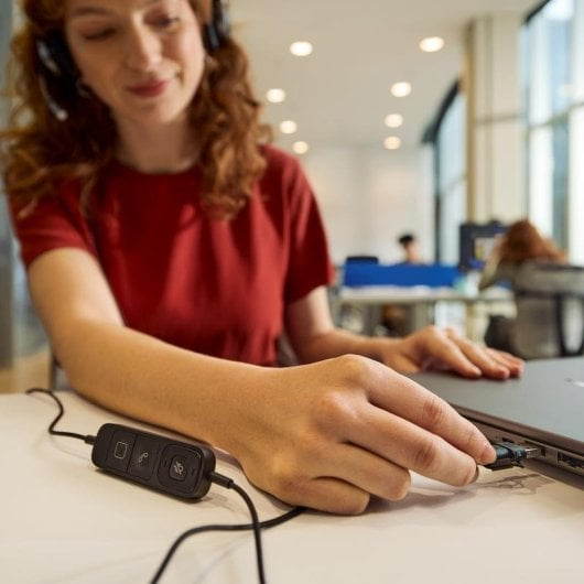Una donna con una maglietta rossa collega le cuffie al laptop in un ufficio.