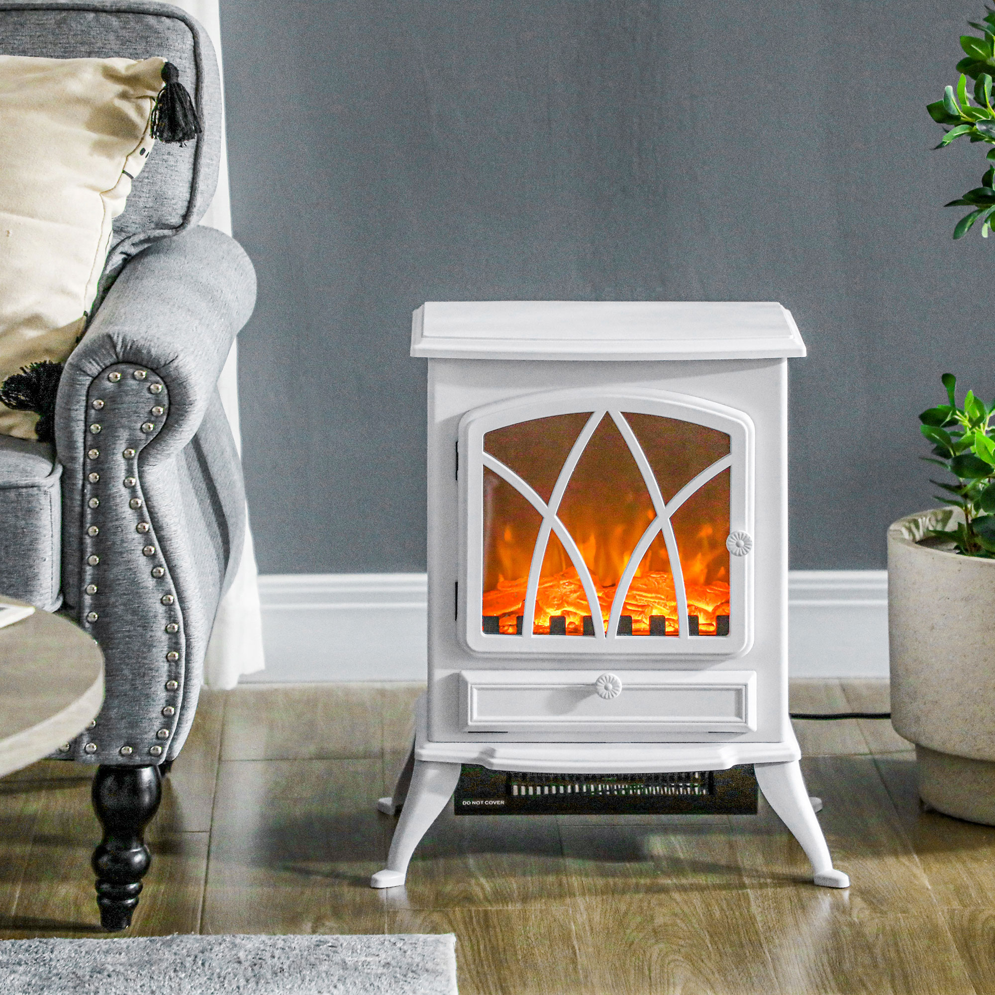 Una chimenea eléctrica blanca sobre un suelo de madera entre un sillón gris y una planta en maceta.