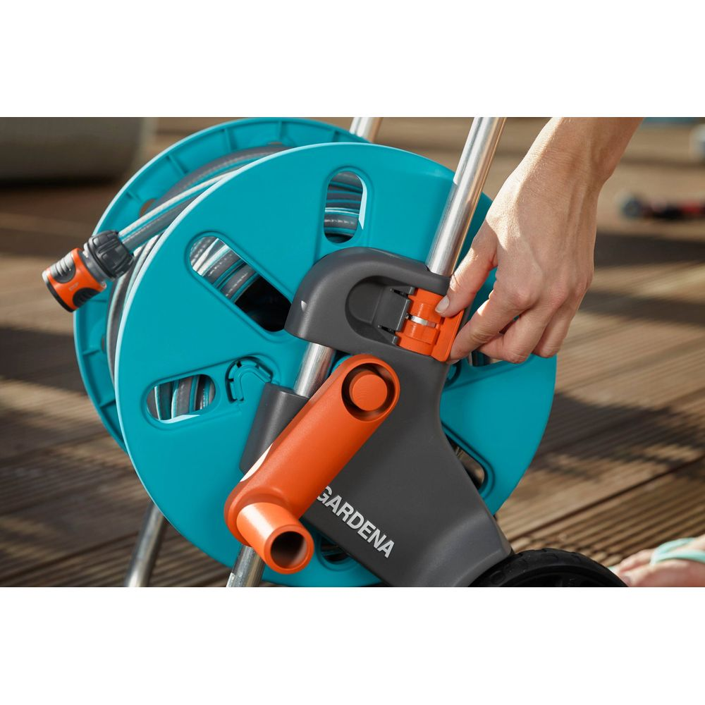 Eine Hand verstellt den orange-grauen Griff eines blauen Gardena Schlauchwagens auf einer Holzterrasse.