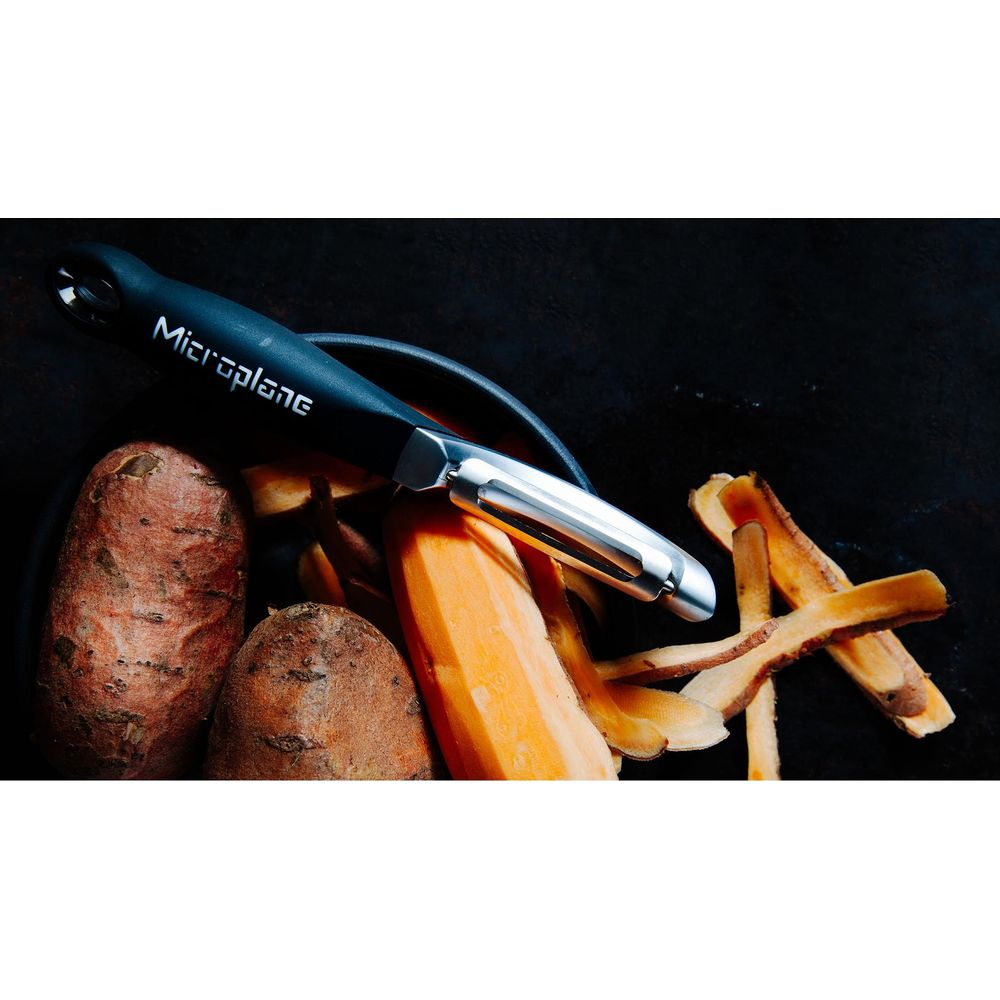 Ein schwarzer Microplane-Sparschäler liegt auf Süßkartoffeln und Gemüseschalen vor dunklem Hintergrund.