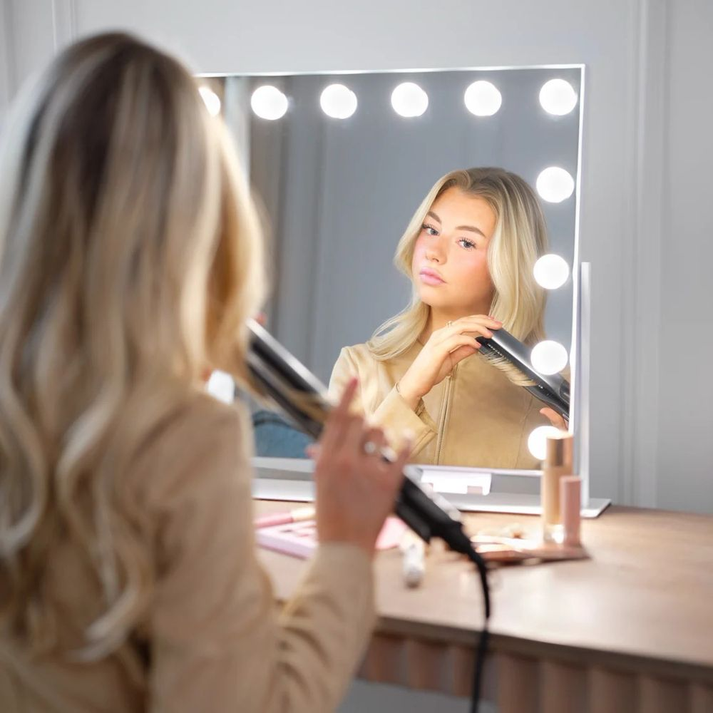 Eine blonde Frau stylt ihre Haare vor einem Leuchtspiegel an einem Holztisch.