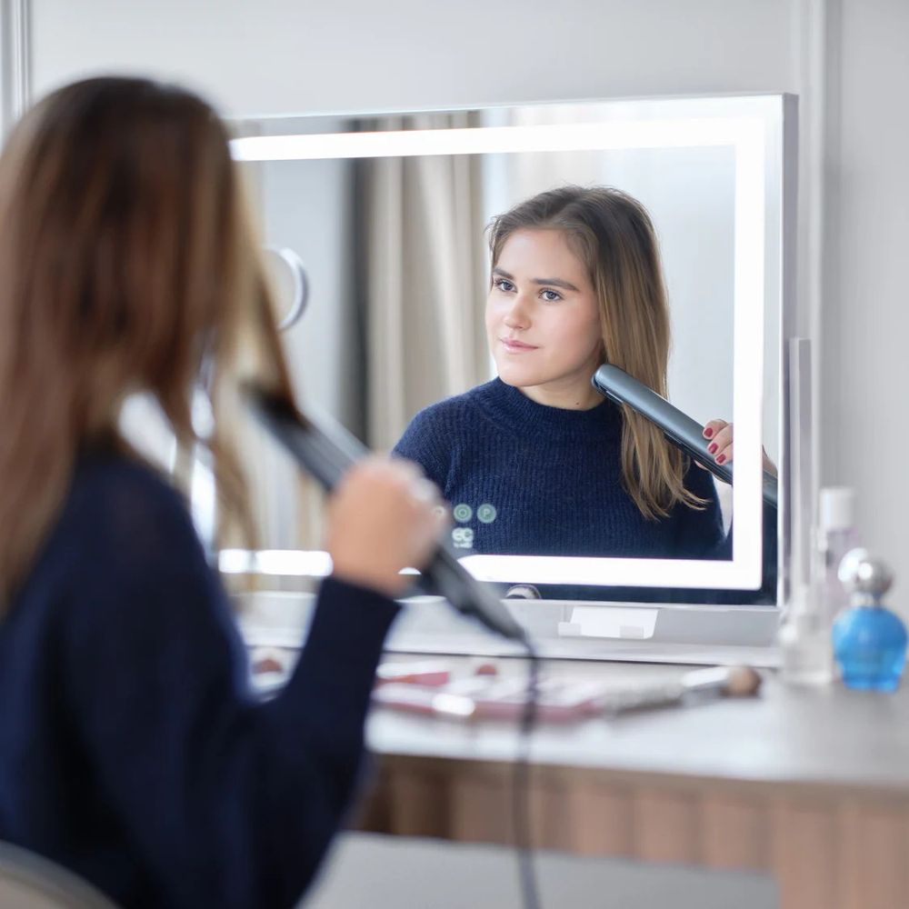 Eine Frau in einem blauen Pullover stylt ihre Haare vor einem beleuchteten Spiegel.