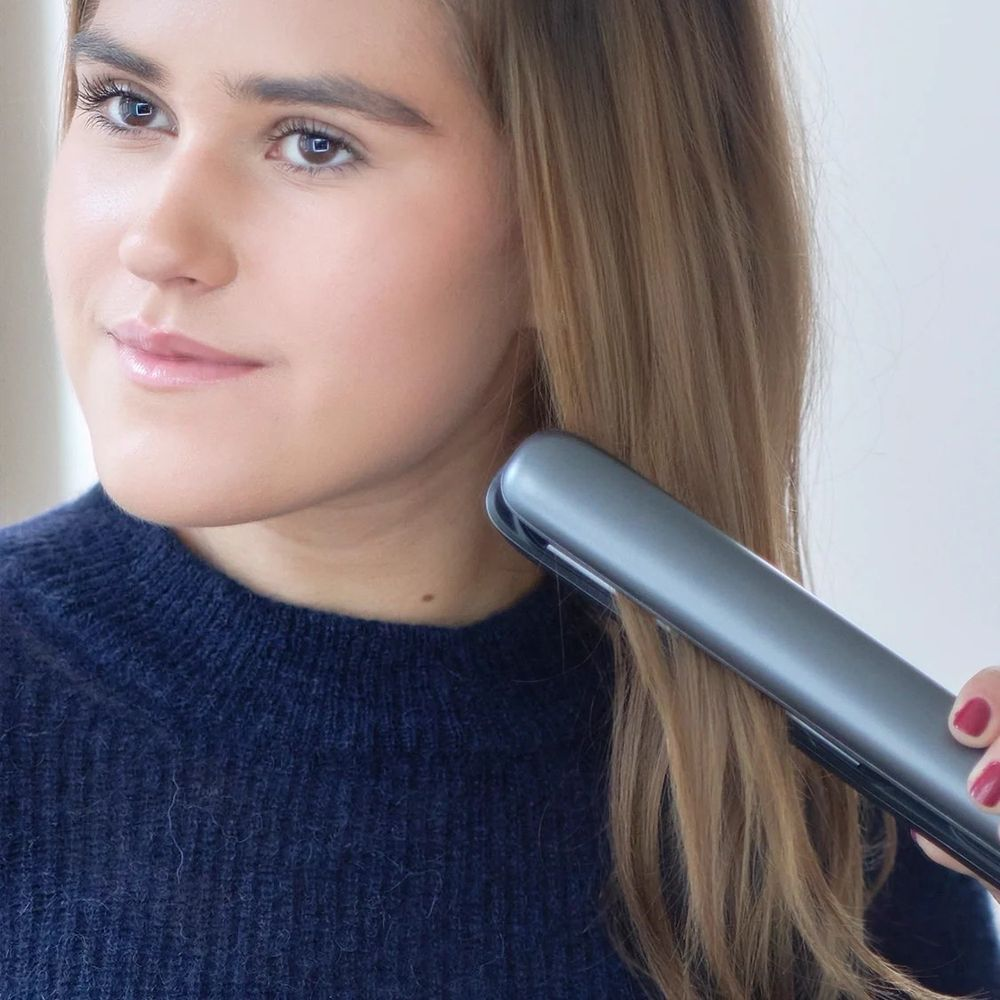Eine Frau in einem blauen Pullover glättet ihre hellbraunen Haare mit einem grauen Haarglättereisen.