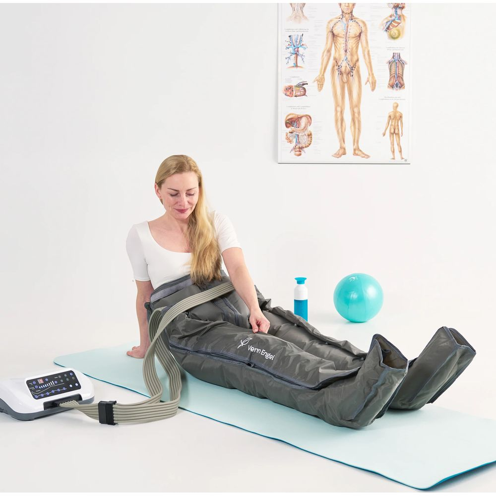 Une femme blonde est assise sur un tapis, portant des bottes de compression grises reliées à un appareil.