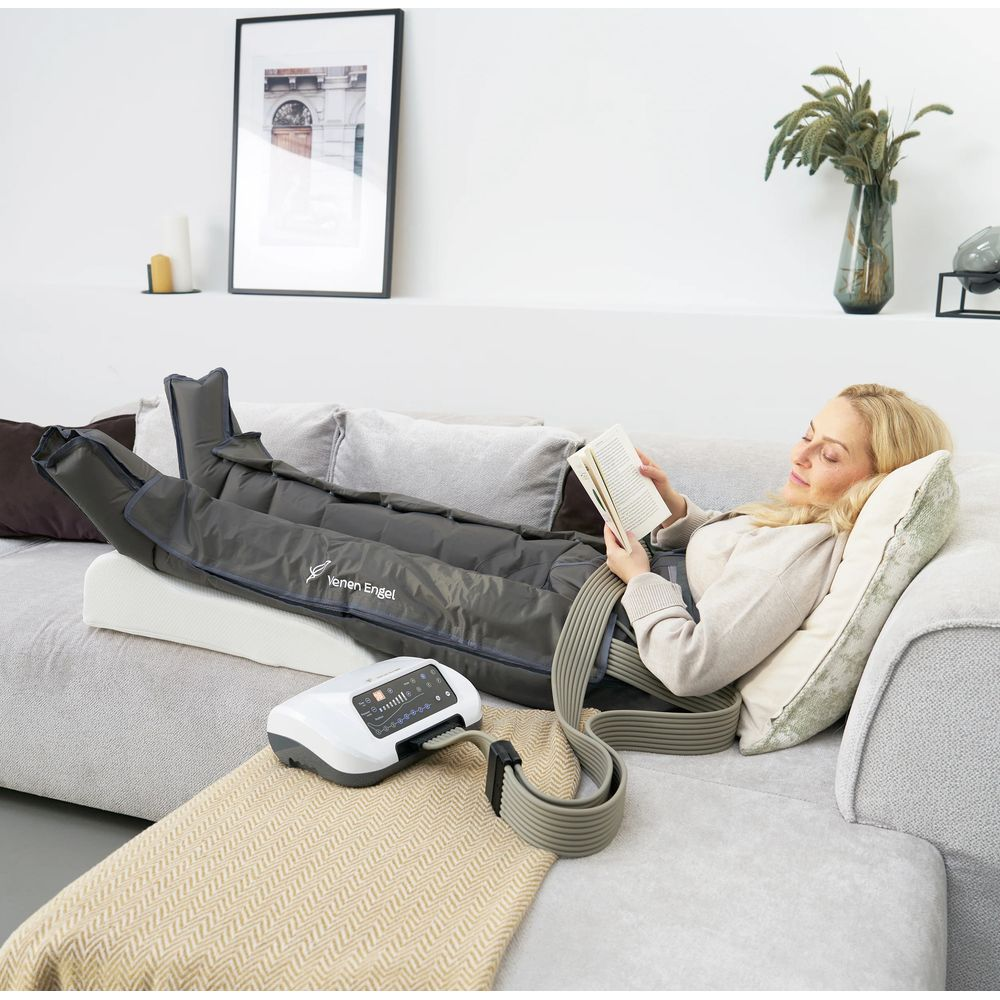 Une femme blonde lit un livre sur un canapé, portant des bottes de compression grises reliées à un boîtier de commande.