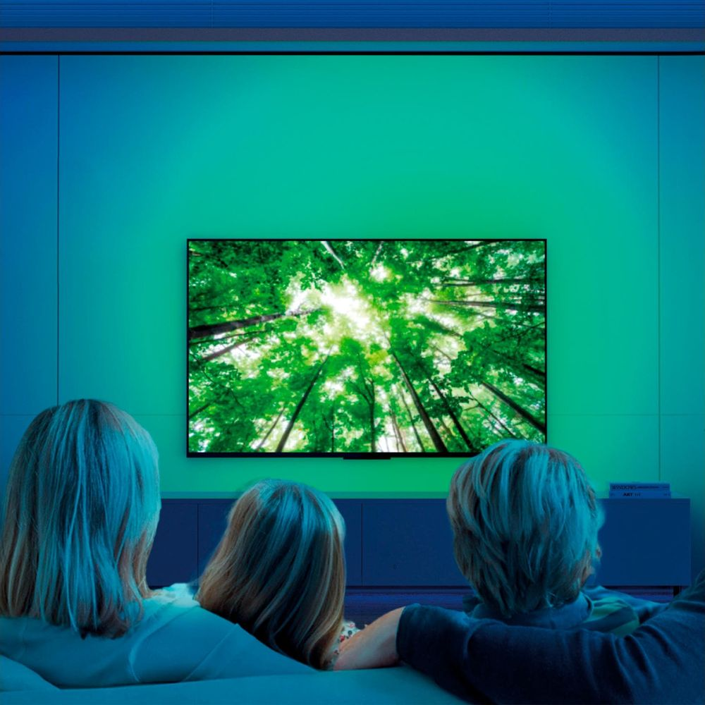 Tres personas vistas de espaldas ven un televisor con árboles verdes, ante una pared con luz ambiental azul verdosa.