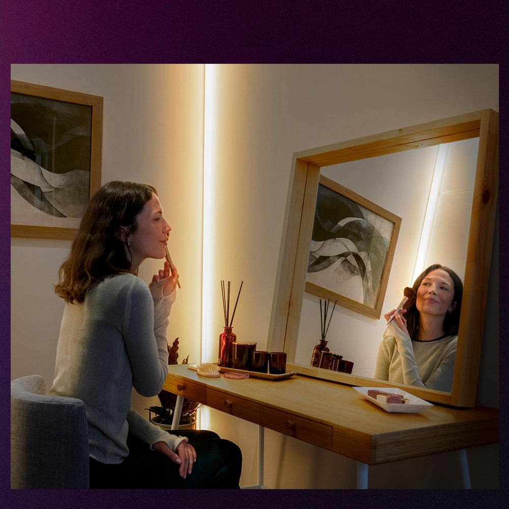 Una donna siede a una toeletta in legno con specchio, truccandosi davanti a una parete illuminata.