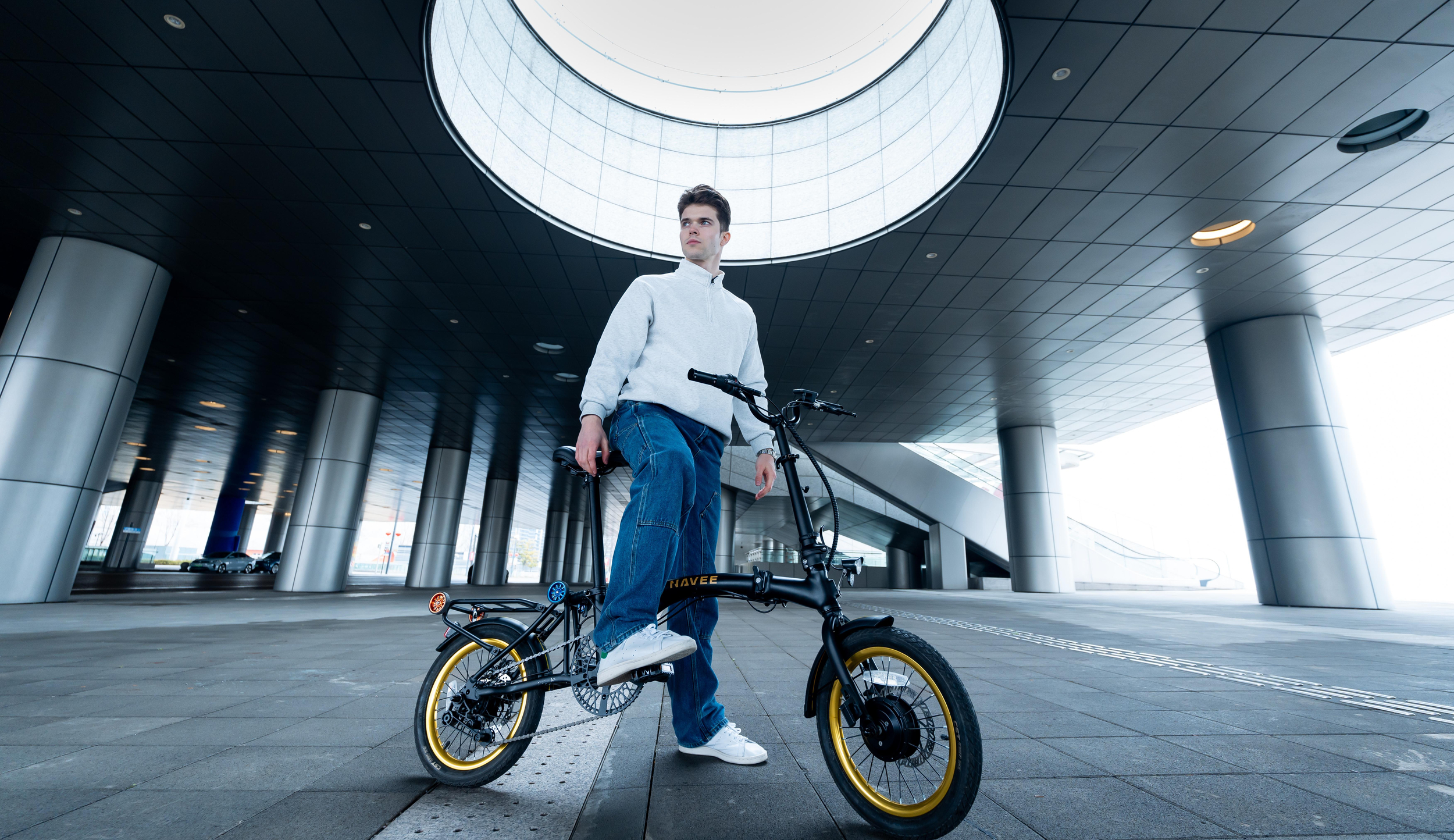 Un giovane con un maglione grigio è in piedi su una e-bike davanti a un'architettura moderna.