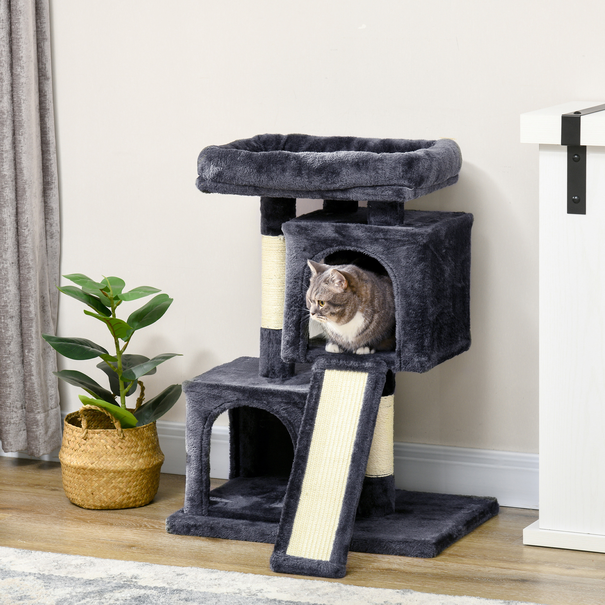 Un gatto grigio e bianco sporge dalla casetta superiore di un tiragraffi grigio, accanto a una pianta verde.