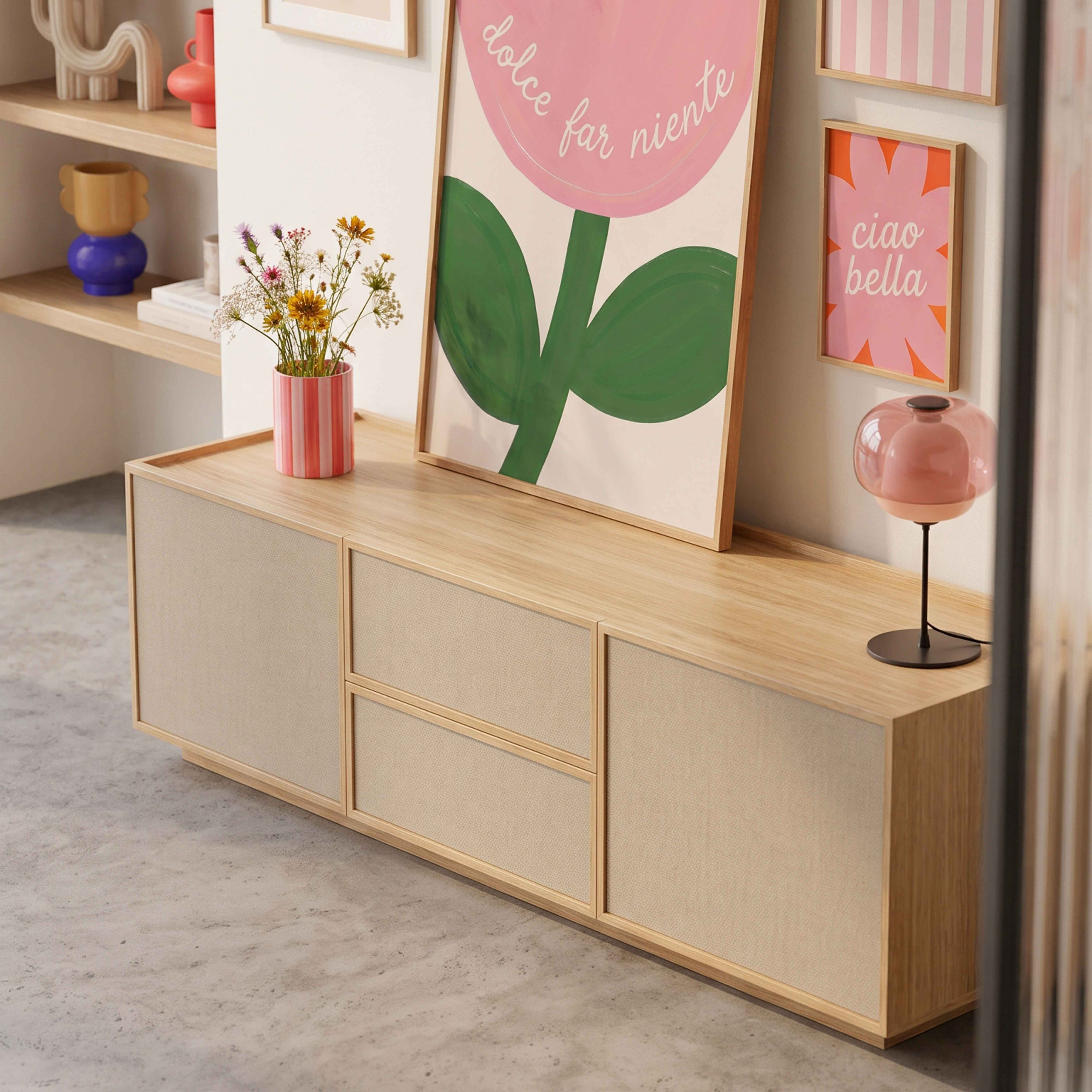 Ein Holzmöbelstück mit beigen Türen, darauf eine Blumenvase, ein gerahmtes Bild und eine rosa Lampe.