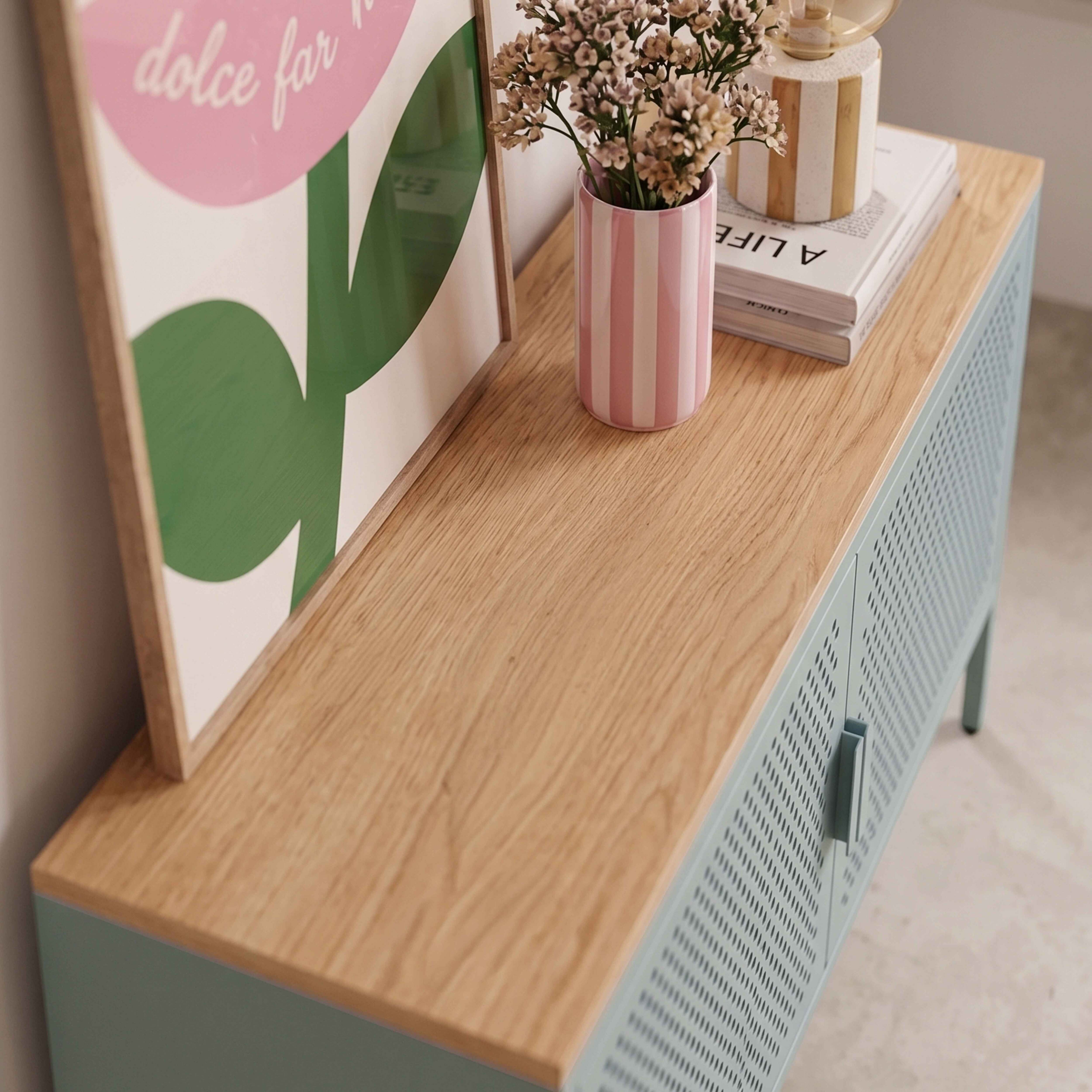 Ein holzfarbener Schrank mit blauen Türen, darauf ein gerahmtes Bild, eine rosa Vase mit Blumen und ein Buch.