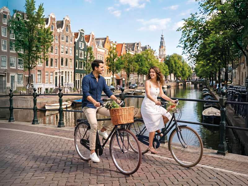 Una pareja sonriente pasea en bicicleta junto a un canal adoquinado en Ámsterdam, rodeada de edificios históricos.